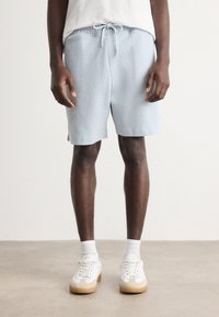 Shorts en tissu côtelé bleu clair avec une ceinture élastique et un cordon de serrage ajustable. Portés avec des baskets blanches et des chaussettes montantes.