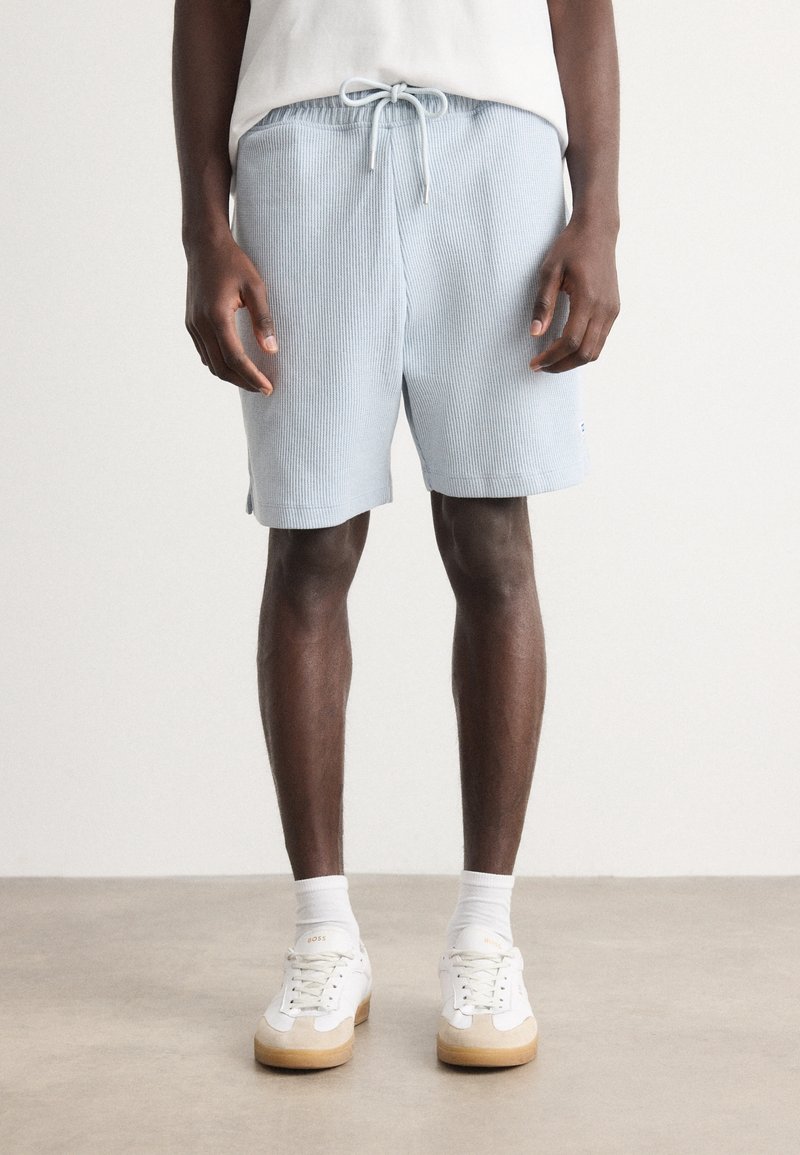 Shorts en tissu côtelé bleu clair avec une ceinture élastique et un cordon de serrage ajustable. Portés avec des baskets blanches et des chaussettes montantes.