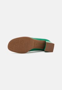 Chaussure à talon vert avec un dessus en cuir lisse et un talon épais. La semelle présente un texte en relief indiquant "Fabriqué avec amour en Espagne."