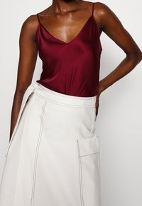 Bordeaux satijnen topje met dunne spaghettibandjes, gecombineerd met een crèmekleurige rok met een striktaille en zijzakken, benadrukt door contrasterende stiksels.