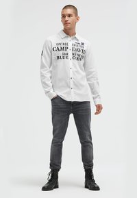 Weißes Langarmhemd mit schwarzen Textgrafiken; Knopfleiste-Design, runder Kragen, unter grauen Jeans und schwarzen Stiefeln geschichtet.