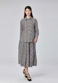 Femme portant un chemisier à boutons noir et blanc à motifs floraux avec une jupe midi assortie et des escarpins bas noirs à lanières, se tenant sur un fond gris clair.