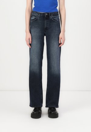 High-waisted, straight-leg jeans in dark blue denim. Features five pockets and a metal button closure. Worn with black platform shoes.
