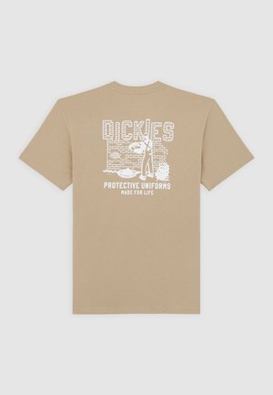 Beige bomulls t-shirt med ett vitt grafiskt tryck av "Dickies Protective Uniforms Made for Life" mot en tegelväggsdesign. Kortsleeve.