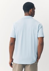 Šviesiai mėlyna polo marškinėlių su lygiu paviršiumi, trumpomis rankovėmis ir klasikiniu apykakle. Nugarinė dalis turi subtilų apvalų kraštą ir minimalų dekorą.