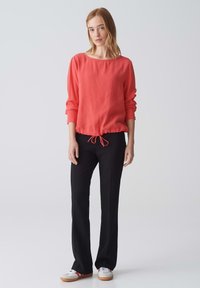 Blusa corallo con scollo rotondo e orlo con coulisse, abbinata a pantaloni neri e sneakers bianche con dettagli rossi. Sfondo semplice.