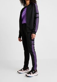 Svart atletisk träningsdräkt med lila ränder, bestående av en zip-jacka och matchande byxor. Sneakers har en svart och rosa färgschema.