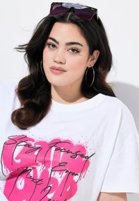 Camiseta blanca de gran tamaño con texto en estilo graffiti en color rosa. La modelo tiene el pelo negro, grandes pendientes de aro dorados y unas gafas de sol reposando sobre su cabeza.