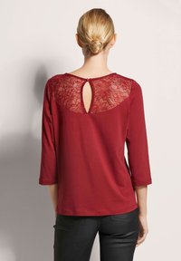 Bordeauxrood overhemd met een kanten inzetstuk, driekwartmouwen en een kleine sleutelgatopening aan de achterkant, afgewerkt met een rood knoopaccent.