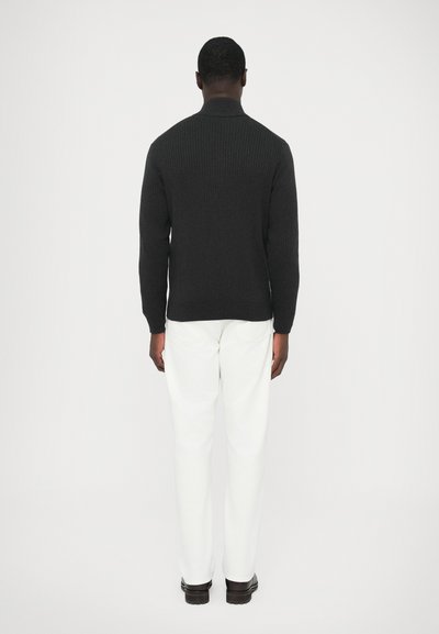 Sötétszürke kötött pulóver magas gallérral, hátulnézetből, fehér nadrággal és fekete cipővel párosítva. Látható a texturált anyag részlete.