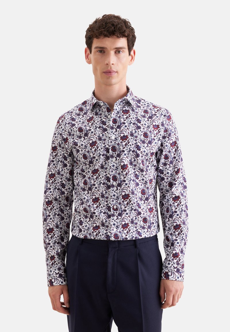 Homme portant une chemise ajustée à manches longues à imprimé floral blanc, violet et rouge, associée à un pantalon bleu marine foncé.