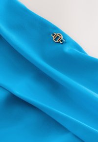 Tissu bleu vif avec des plis lisses et un petit emblème rond en or présentant un motif géométrique cousu près du bord.