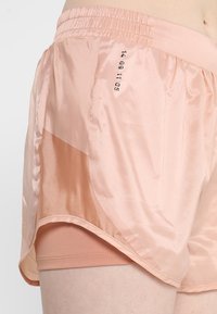 Vaaleanpunaiset juoksushortsit, joissa on satiinipinta, joustava vyötärö ja läpinäkyvä päällyste. Sivulla on painetut numerot.