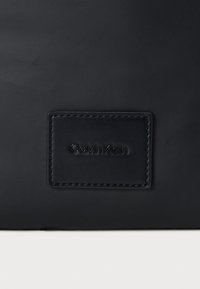 Crna kožna torba s teksturiranom površinom i utisnutim Calvin Klein logotipom na pravokutnom ok patchu. Minimalistički dizajn.