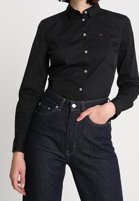 Černá košile s dlouhými rukávy a límcem na knoflíky, s bílými knoflíky a malým logem. Kombinováno s tmavými džíny s vysokým pasem.