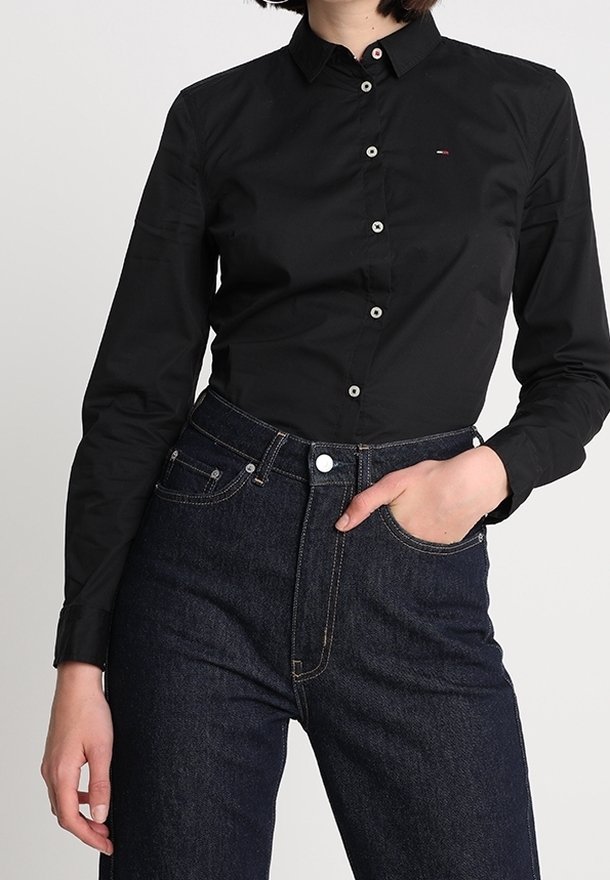 Černá košile s dlouhými rukávy a límcem na knoflíky, s bílými knoflíky a malým logem. Kombinováno s tmavými džíny s vysokým pasem.