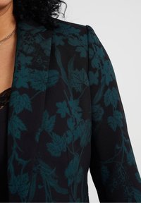 Svart kavaj med ett blommönster i mörk teal. Har strukturerade axlar och slät tygkvalitet, som visar upp intrikata bladdetaljer.