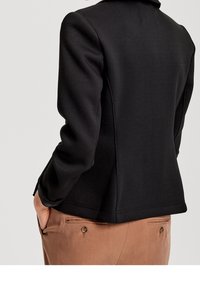 Svart kavaj i texturerad tyg, med skräddarsydda axlar och en figursydd design, i kombination med bruna byxor.