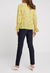 Blouse florale jaune à manches longues et poignets à volants, associée à un pantalon slim-fit bleu marine et des chaussures à talons blanches.