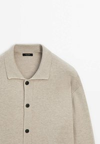 Beige gebreide cardigan met een geribbelde kraag, vijf zwarte knopen en een zachte textuur. Heeft een iets oversized pasvorm en een ontspannen ontwerp.