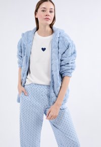 Mujer vistiendo una chaqueta esponjosa azul clara, una camiseta blanca con un pequeño corazón oscuro y pantalones azul claros con estampado, de pie con una mano en el bolsillo.