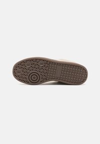 Brown sneaker sole with textured rubber grip, circular pattern details, and a logo embossed on the side, featuring a wavy tread pattern.