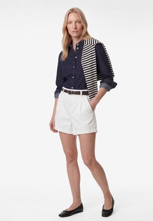 Vrouw met een marineblauw overhemd met knopen, witte short met riem, zwarte platte schoenen, en een zwart-wit gestreepte trui over haar schouders gedrapeerd.