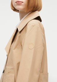 Άτομο με κοντά κόκκινα μαλλιά που φοράει ένα μπεζ παλτό τύπου trench με στρογγυλή μπλε επένδυση λογότυπου στον πάνω βραχίονα, σε έναν απλό φόντο.