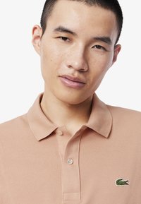 Polo-paita vaalean persikan värissä, valmistettu teksturoituneesta kankaasta. Ominaisuuksina klassinen kaulus, nappilista ja pieni vihreä Lacoste-logo rinnassa.