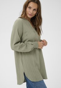 Cămașă cu mânecă lungă de culoare verde deschis, confecționată dintr-un material texturat cu dungi verticale, având decolteu în V și manșete adunate. Dispune de deschideri laterale.