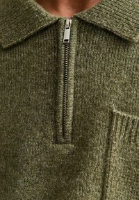 Suéter de punto en verde olivo con un cuello con cremallera. Presenta una textura acanalada y un pequeño bolsillo en el lado izquierdo. Fabricado en una tela suave y gruesa.