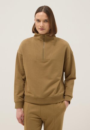 Frau trägt einen braunen Pullover mit einem halben Reißverschluss am Kragen und passende Hose, steht mit einer Hand in der Tasche vor einem einfarbigen Hintergrund.