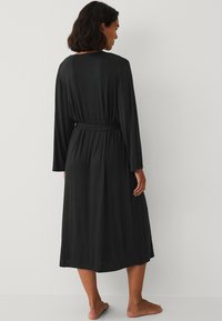 Robe noire à manches longues en tissu doux, dotée d'une taille nouée et d'un design fluide, atteignant le mollet avec une texture lisse.