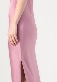 Robe en satin rose avec une silhouette ajustée et une fente sur le côté, mettant en valeur une texture lisse et un subtil éclat. Main reposant sur le tissu.