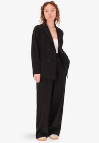 Blazer nero doppio petto con ampie revers, abbinato a pantaloni larghi coordinati e una maglietta bianca; il modello indossa scarpe chiare.