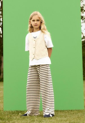 Beige Leinenweste über einem weißen T-Shirt, kombiniert mit gestreiften, weiten Hosen. Blaue Schuhe auf Gras, vor einem grünen Hintergrund.