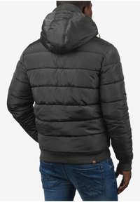 Gepolsterte schwarze Jacke mit Kapuze, horizontal gesteppten Abschnitten und gerippten Bündchen. Dazu blaue Jeans mit strukturiertem Design.