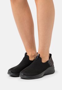 Black slip-on shoes with a knitted upper, featuring a textured finish and a rounded toe. Dark gray accents at the heel provide contrast.