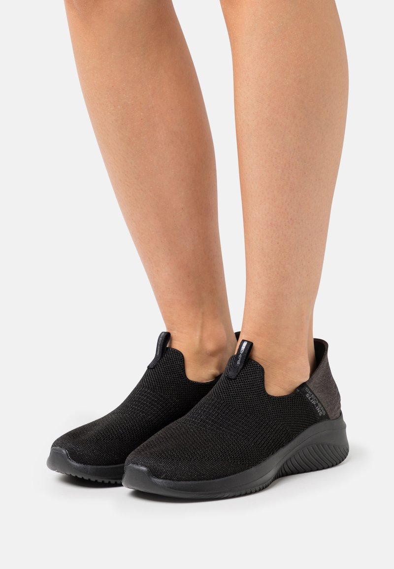 Black slip-on shoes with a knitted upper, featuring a textured finish and a rounded toe. Dark gray accents at the heel provide contrast.