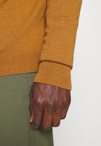 Maglione lavorato a maglia color senape con polsini a coste, abbinato a pantaloni verde oliva. Primo piano di una mano contro il tessuto testurizzato.