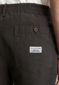 Donkergroene elastische broek met een achterzak met een bruine knop en een wit label met de tekst "Marc O'Polo Denim." Glad stoffen textuur.