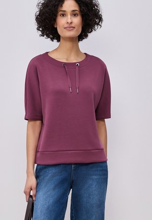 Femme aux cheveux bouclés portant un haut bordeaux à manches courtes avec des cordons, un jean bleu et tenant un sac à main sombre à côté d'elle.