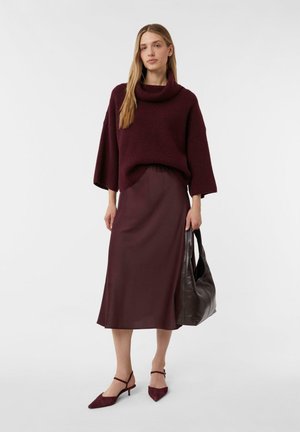 Frau mit einem burgunderfarbenen Rollkragenpullover, einem passenden Midi-Rock, burgunderfarbenen spitzen High Heels und einer braunen Lederhandtasche.