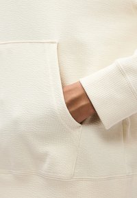 Χέρι με σκούρο δέρμα εισέρχεται στην εμπρόσθια τσέπη μανικιού υφασμάτινης, κρεμ χρωματισμένης, υφής που μοιάζει με πουλόβερ ή φούτερ.