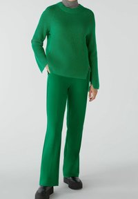 Pull en maille côtelée verte avec des manches longues et un détail de zip sur le côté, associé à un pantalon vert ajusté, porté avec des chaussures à plateforme noires.