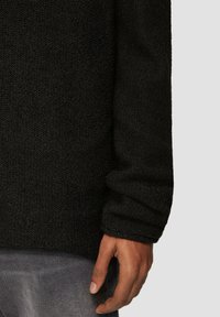 Schwarzer strukturierter Pullover mit lockerer Passform und langen Ärmeln, versehen mit einem gerippten Bündchen. Hand sichtbar auf grauem Stoff ruhend.