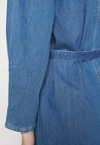 Casaco de ganga com cintura cingida, mangas longas com punhos verdadeados e uma cor azul desbotada com detalhes de costura visíveis.