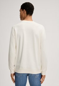 Kremowy sweterek z okrągłym dekoltem, długimi rękawami, prążkowanymi mankietami i dołem. Gładka faktura, luźny krój, noszony z niebieskimi dżinsami.