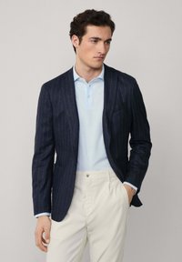Námořnický blazer s jemným proužkem a upraveným střihem, vyrobený z texturované látky. Nosený přes světle modrou polokošili a krémové kalhoty.