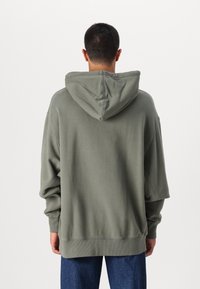 Felpa verde oversize con cappuccio regolabile, polsini e orlo a coste, caratterizzata da un tessuto liscio e testurizzato. Vista posteriore.